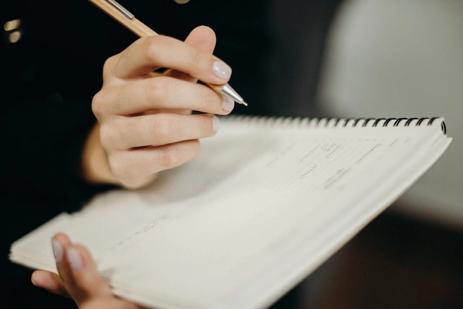 Osoba si zapisuje poznámky do zápisníku u stolu s notebookem